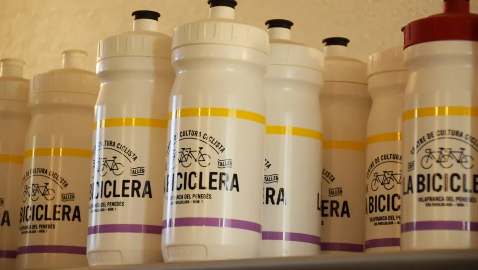 Bidons blancs serigrafiats amb el nom de La Biciclera, alineats en una prestatgeria.